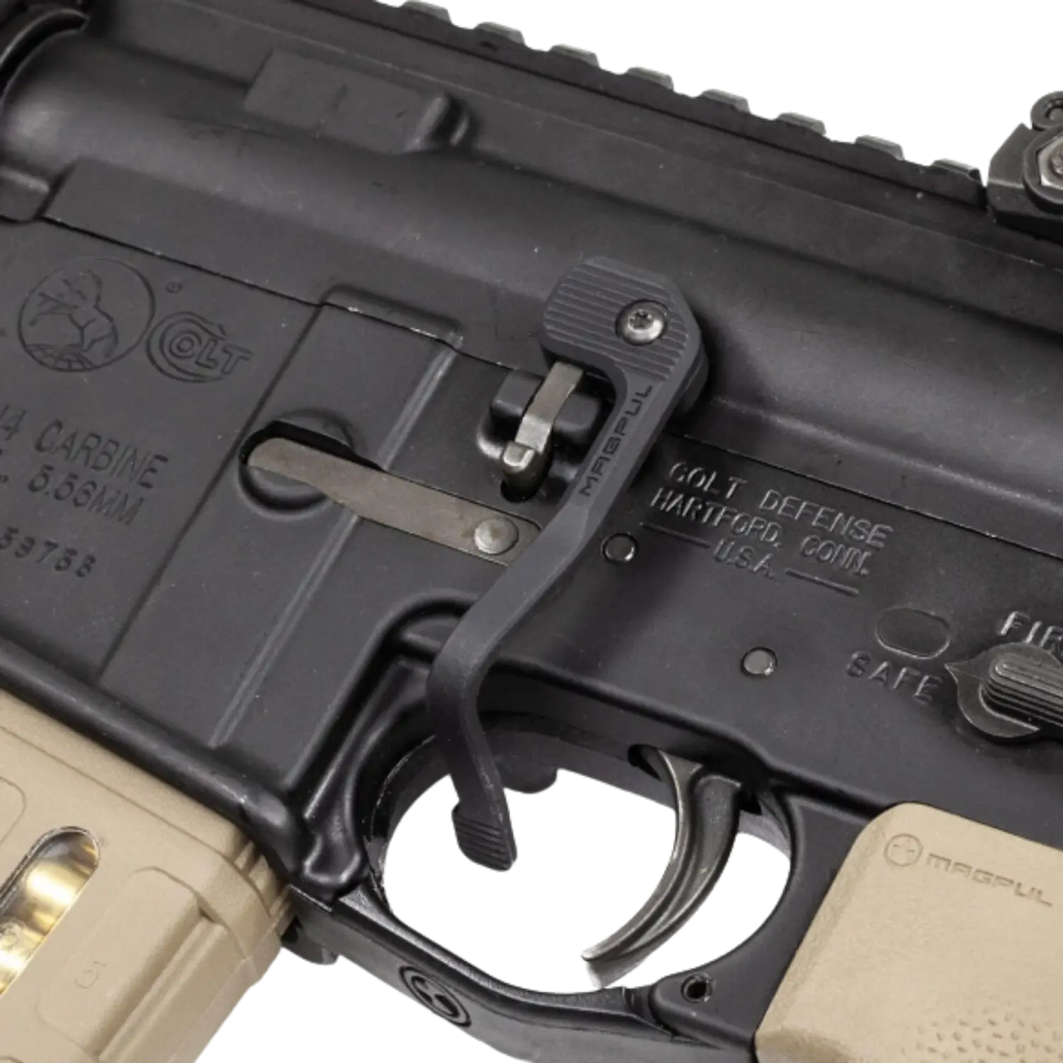 Nahaufnahme eines schwarzen MAGLULA B.A.D. Battery Assist Device Hebels am Abzugsbügel eines AR-15/M4-Gewehrs mit sichtbaren Gravuren und beigem Griffstück.